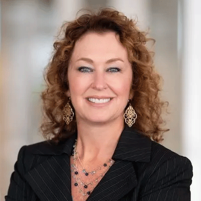 Marybeth Moran - Texas Hospital Association Head-and-shoulders portrait of a smiling woman with curly red hair, wearing a black pinstripe blazer and gold dangling earrings.