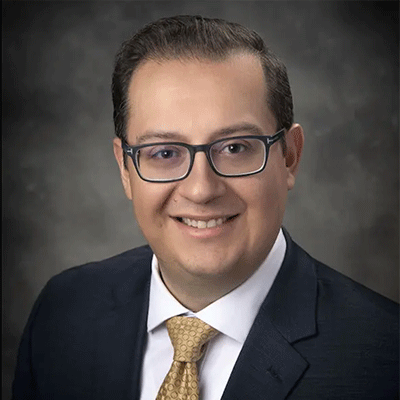 Oliver Ostorga - Texas Hospital Association Smiling man in a dark suit and yellow tie, wearing rectangular glasses, studio portrait with a gray backdrop.