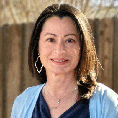 Silvia Rose - Texas Hospital Association Smiling woman with shoulder-length dark hair wearing a blue cardigan and hoop earrings, outdoors with a wooden fence in the background.
