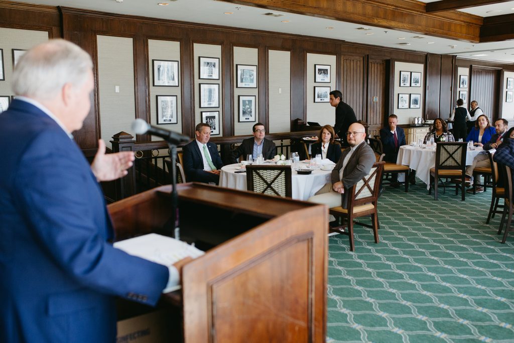 07242026_Fellows_ModIV_nguillot-45 - Texas Hospital Association Speaker at a wooden lectern addresses a formal luncheon audience, seated at round tables in a paneled room with framed photos on the walls (foreground man blurred).