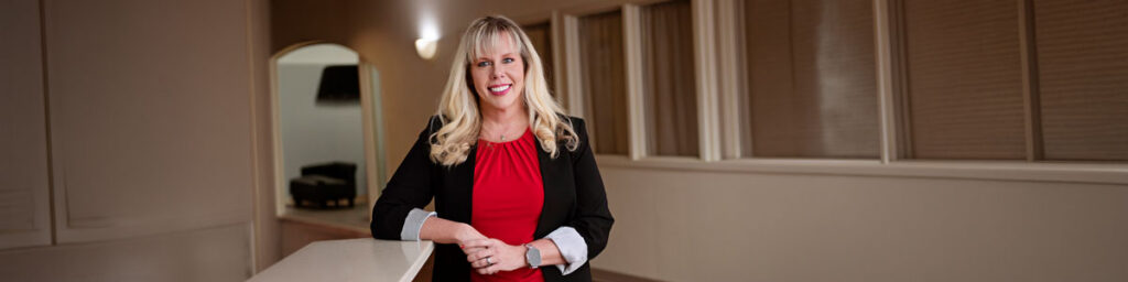 Candice Smith, 2026 Pioneer Award Winner - Texas Hospital Association Professional woman in a black blazer and red top standing at a light counter in an office hallway, smiling at the camera.