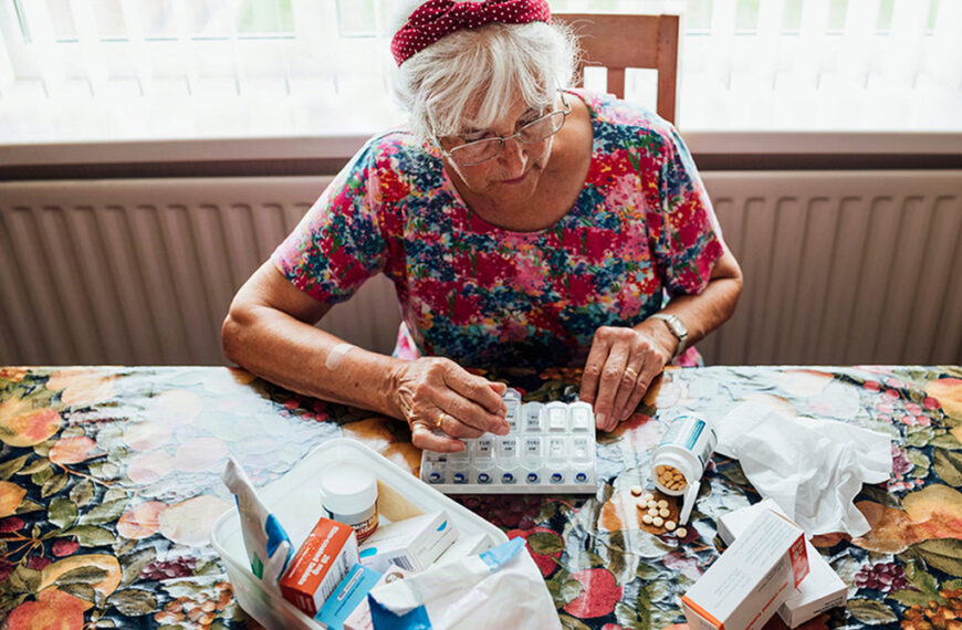 Elderly woman in a floral shirt sorting pills into a weekly pill organizer at a table full of medications and boxes.