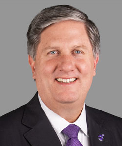 Smiling, middle-aged man in a dark suit and purple tie, head-and-shoulders portrait on a gray background; visible lapel pin.