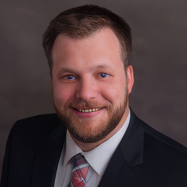 Professional headshot of a smiling man in a dark suit and pink striped tie against a neutral backdrop.