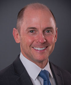 Professional headshot of a smiling middle-aged man in a dark suit and blue tie, gray studio background.