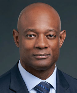 Professional headshot of a bald Black man in a navy suit and blue tie, looking at the camera.