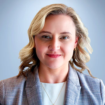 Professional headshot of a smiling woman with blonde hair in a light blazer against a blue background.