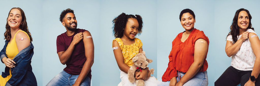 Five diverse adults smile at the camera, each with a bandage on their upper arm after vaccination against a light blue background.