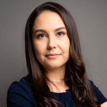 Portrait of a woman with long dark hair wearing a navy blouse, looking at the camera against a gray studio background.
