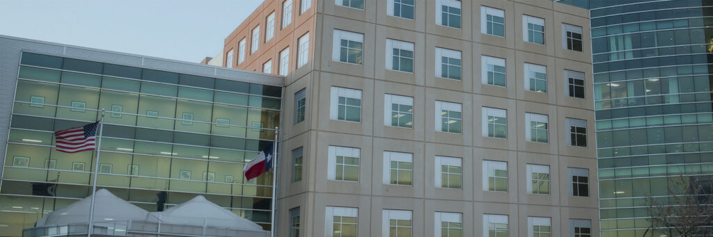 Modern glass-fronted office building with a beige brick facade and US and Texas flags at the entrance.