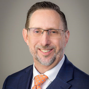 Professional headshot: smiling man in a navy suit, white shirt, orange polka-dot tie, wearing glasses.