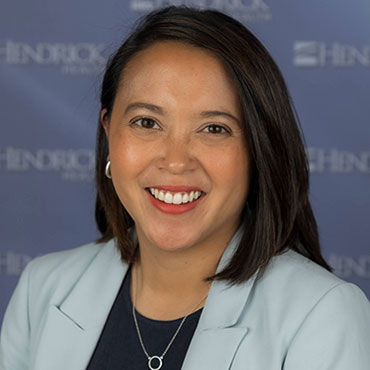 Professional headshot of a smiling woman with shoulder-length dark hair in a light blue blazer against a blue backdrop.