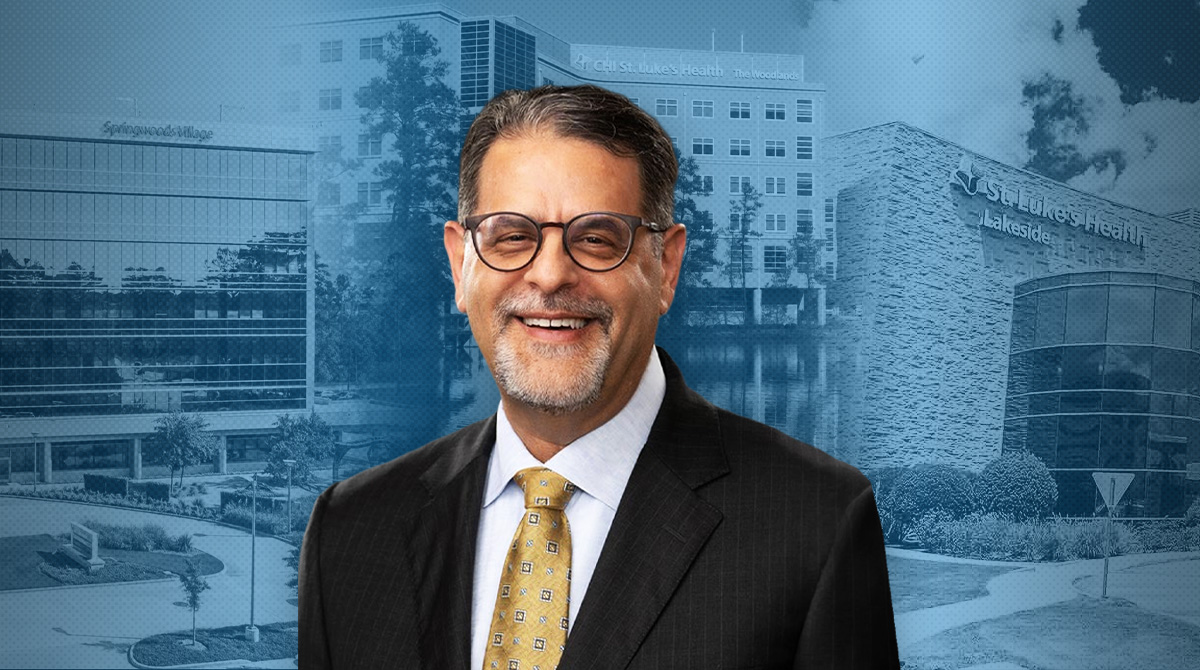 Smiling middle-aged man in a dark suit and yellow tie stands in front of a blue-tinted medical campus backdrop.