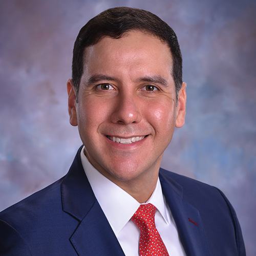 Jorge Leal - Texas Hospital Association Head-and-shoulders portrait of a smiling man in a navy suit with a red tie against a soft blue studio background