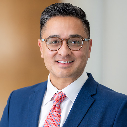 Joshua Pozos - Texas Hospital Association Portrait of a smiling man in a blue suit, white shirt, pink striped tie, and glasses in an office setting.