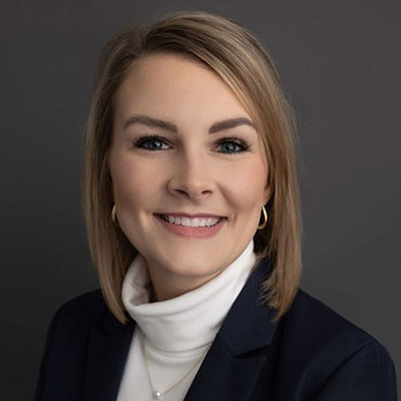 Professional headshot: smiling woman in a navy blazer, white turtleneck, against a gray background.
