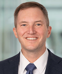 Portrait of a smiling man in a navy suit and blue tie, in a neutral office backdrop.
