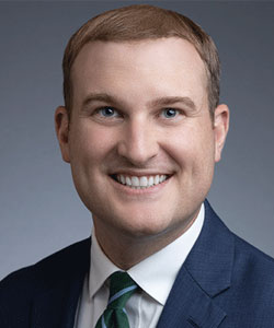 Professional headshot: smiling man in a navy suit and green tie against a gray backdrop.