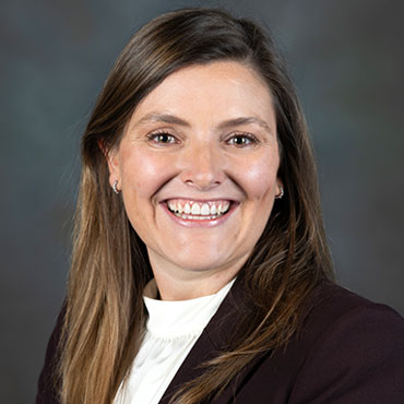 Professional headshot of a smiling woman with long brown hair, wearing a white blouse and dark blazer against a muted backdrop.