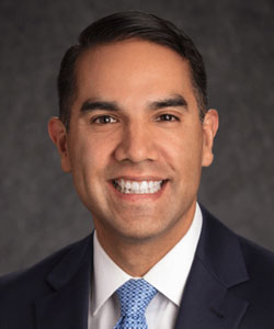 Head-and-shoulders portrait of a smiling man in a dark suit and light blue tie against a gray backdrop.