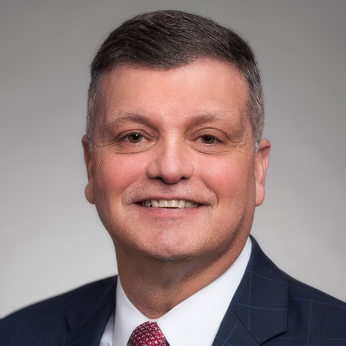 Noel Cárdenas - Texas Hospital Association Smiling man in a navy suit and red tie, head-and-shoulders portrait against a neutral gray background for a professional headshot.