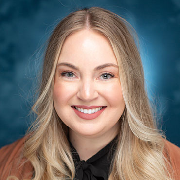 Portrait of a smiling woman with long blonde hair, wearing a black blouse and brown jacket, against a blue backdrop.