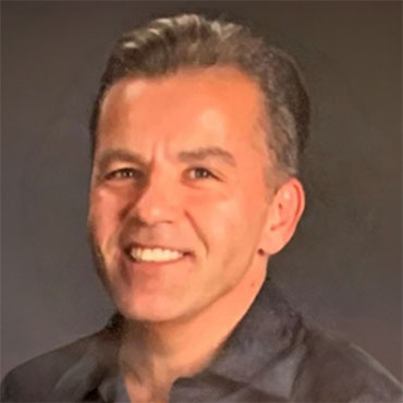 Portrait of a smiling man with short gray hair in a dark shirt, against a dark studio background.