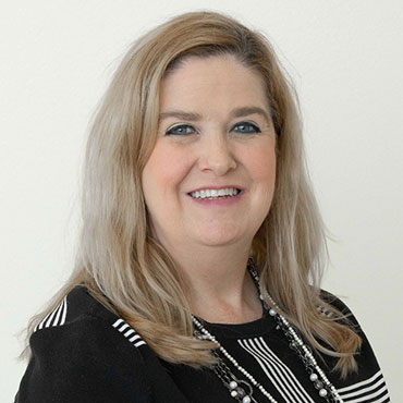Portrait of a smiling woman with shoulder-length blonde hair wearing a black blazer and beaded necklace against a light background, a professional headshot.
