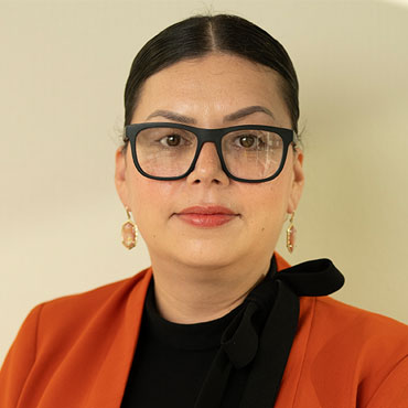 Professional portrait of a woman with dark hair, black-framed glasses, orange blazer, and gold earrings against a light background.