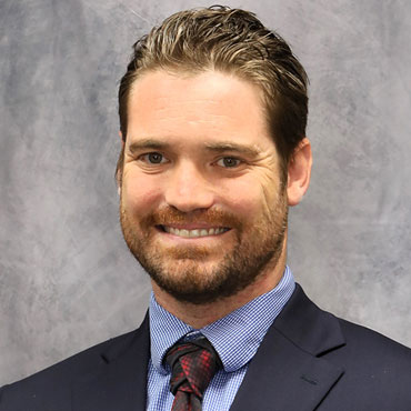 Professional headshot of a smiling man in a navy suit, blue shirt, and red tie against a gray backdrop.
