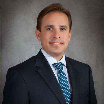 Shane Casady - Texas Hospital Association Professional headshot of a smiling man in a navy suit and blue tie against a gray backdrop for a corporate profile.