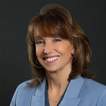 Professional portrait of a smiling woman in a light blue blazer against a dark background, suitable for an About page.