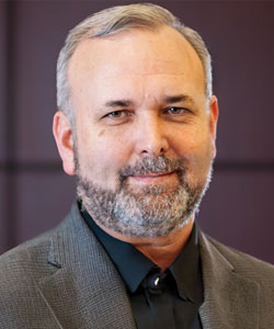 Portrait of a middle-aged man with gray hair and beard, wearing a dark shirt and gray blazer, smiling slightly.
