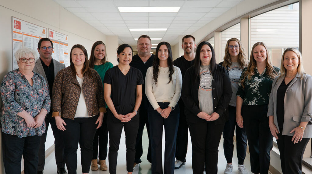 Twelve employees standing in a bright office hallway, facing the camera and smiling, with posters on the wall to the left and large windows to the right beneath a drop ceiling.