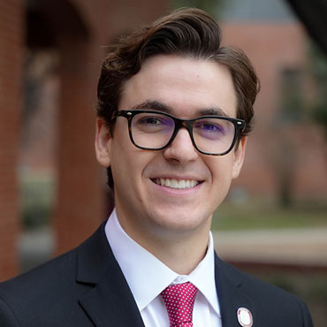Smiling man in a dark suit, white shirt, pink tie, and black-framed glasses outdoors (portrait).