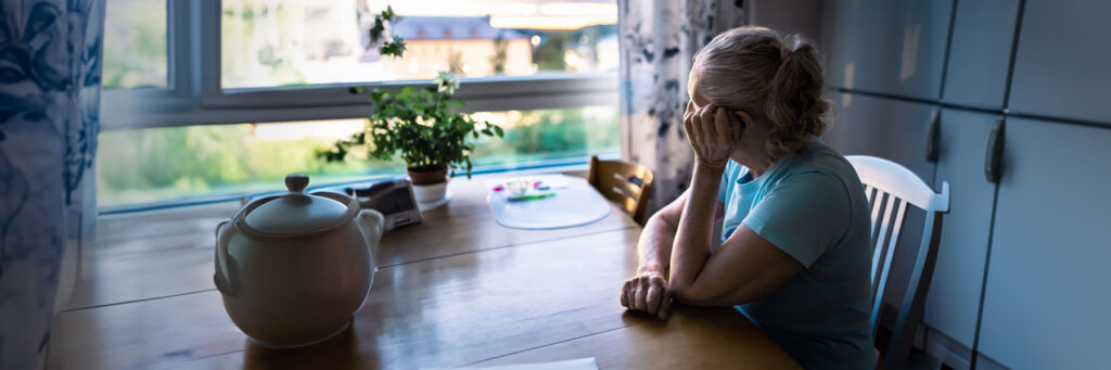 Older woman sits at a kitchen table by a window, head in her hand in a gesture of worry or fatigue.
