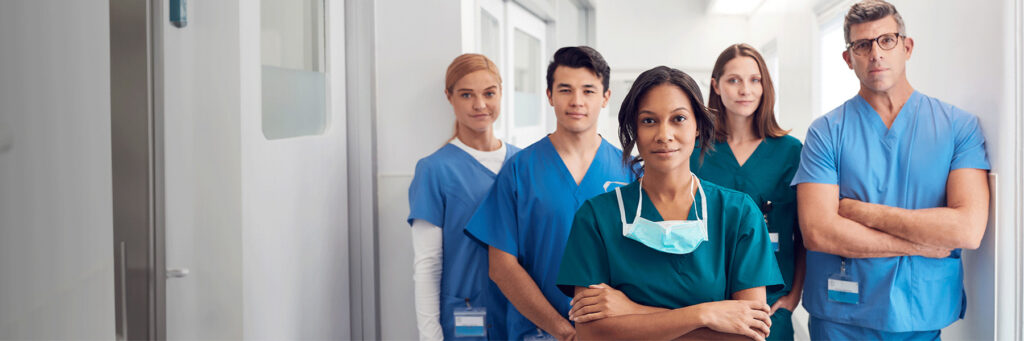Group of diverse medical professionals in scrubs standing in a bright hospital corridor, looking at the camera.