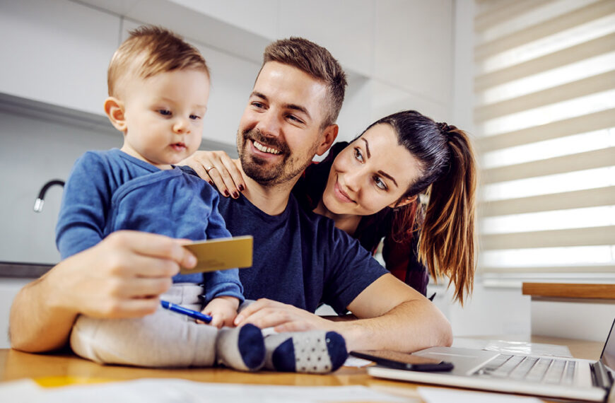 Family of three at a kitchen table looking at a laptop; dad holds a card while the toddler sits on his lap, smiling popular scene of online shopping.