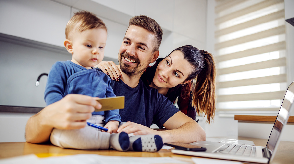 Family of three at a kitchen table looking at a laptop; dad holds a card while the toddler sits on his lap, smiling popular scene of online shopping.